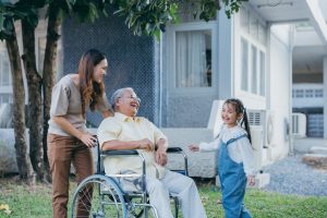caregiver job responsibilities | Disabled senior grandpa on wheelchair with grandchild and mother in park, Happy Asian multi generation family having fun together outdoors backyard, Grandpa elderly and little child smiling and laugh.