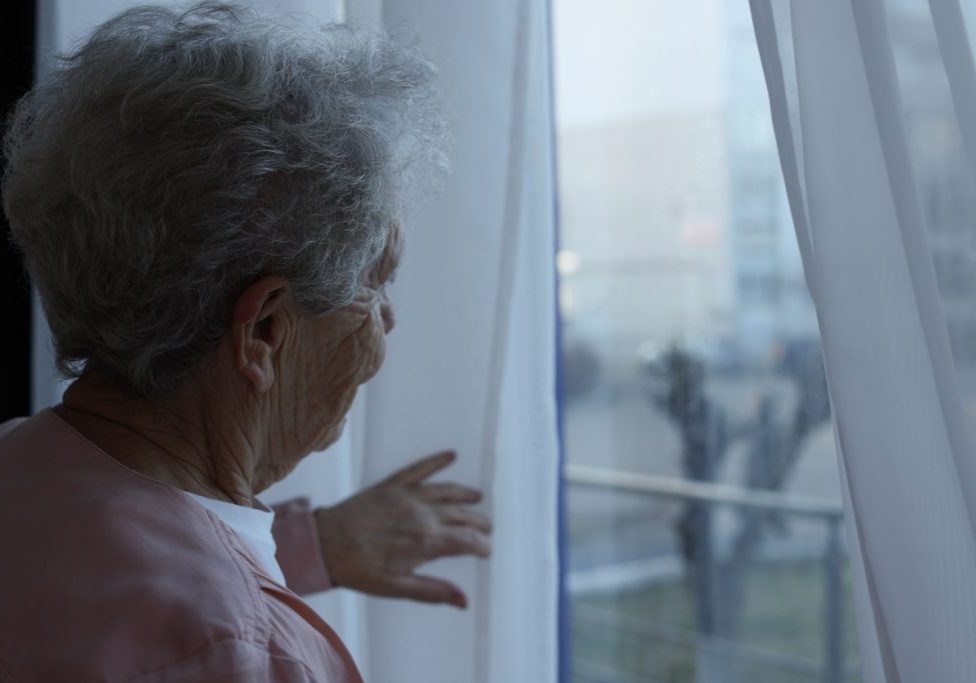 Elderly woman looking out of window indoors, space for text. Loneliness concept