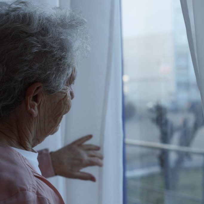 Elderly woman looking out of window indoors, space for text. Loneliness concept